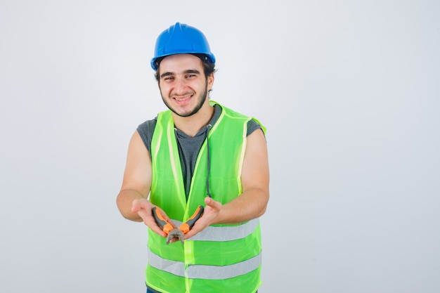 Молодой строитель человек показывает плоскогубцы в форме спецодежды и выглядит радостным, вид спереди.