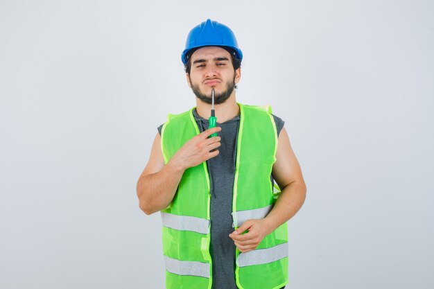 Young builder man in uniform holding tip of screwdriver on chin and looking thoughtful , front view.