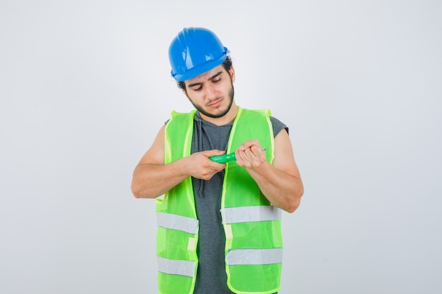 Young builder man in uniform looking at screwdriver and looking thoughtful , front view.