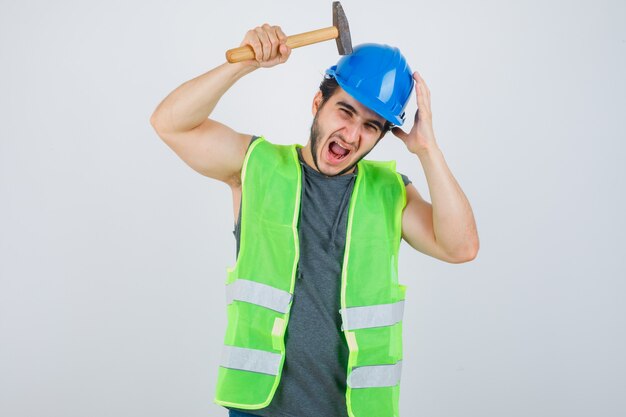 Young builder man in uniform striking head with hammer and looking funny , front view.