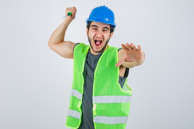 Young builder man in uniform striking with screwdriver and looking crazy , front view.