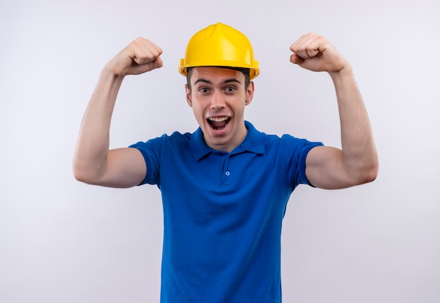 Uomo giovane costruttore indossa uniforme da costruzione e casco di sicurezza facendo pugni felici
