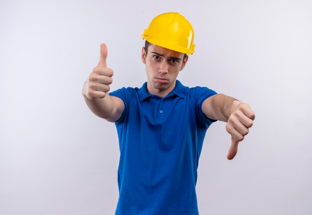 Молодой строитель человек в строительной форме и защитном шлеме делает счастливые пальцы вверх и несчастные пальцы вниз