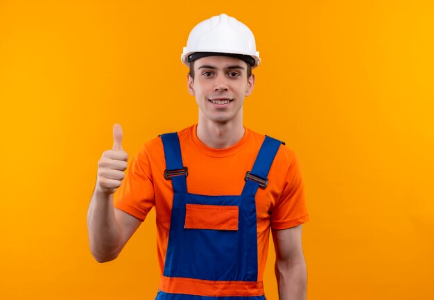 Молодой строитель человек в строительной форме и защитном шлеме делает счастливые пальцы вверх