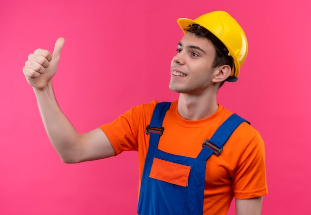 Uomo del giovane costruttore che indossa l'uniforme della costruzione e il casco di sicurezza che fa i pollici felici in su