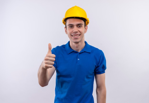 Молодой строитель человек в строительной форме и защитном шлеме делает счастливые пальцы вверх