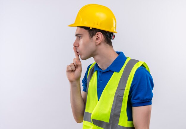 Young builder man wearing construction uniform and safety helmet doing silence gesture with finger