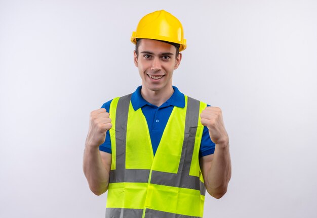 Young builder man wearing construction uniform and safety helmet happily shows power with fists