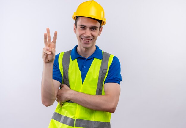 건설 유니폼과 안전 헬멧을 착용하는 젊은 작성기 남자는 행복하게 세 손가락을 보여줍니다