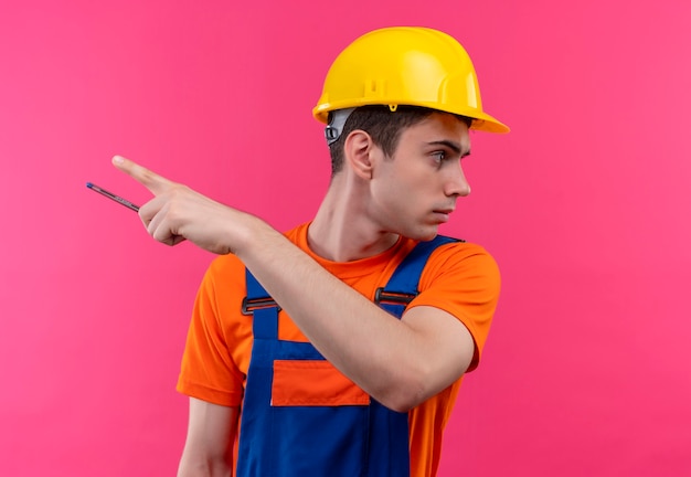 Young builder man wearing construction uniform and safety helmet holds a pen and points to the left with thumb