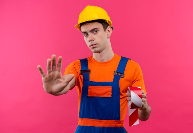 건설 유니폼과 안전 헬멧을 착용하는 젊은 작성기 남자는 빨간색 흰색 신호를 보유하고 그의 손으로 중지를 보여줍니다