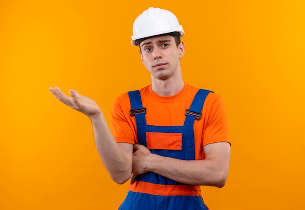 Молодой строитель в строительной форме и защитном шлеме держит левую руку вверх