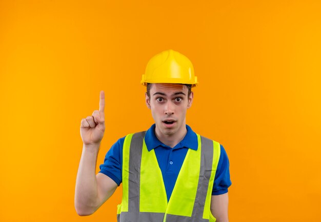 Young builder man wearing construction uniform and safety helmet point up with finger suprised