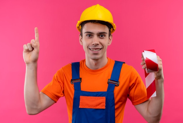 건설 유니폼 및 안전 헬멧을 착용하는 젊은 작성기 남자 엄지 손가락을 가리키고 빨강-흰색 신호 테이프를 보유