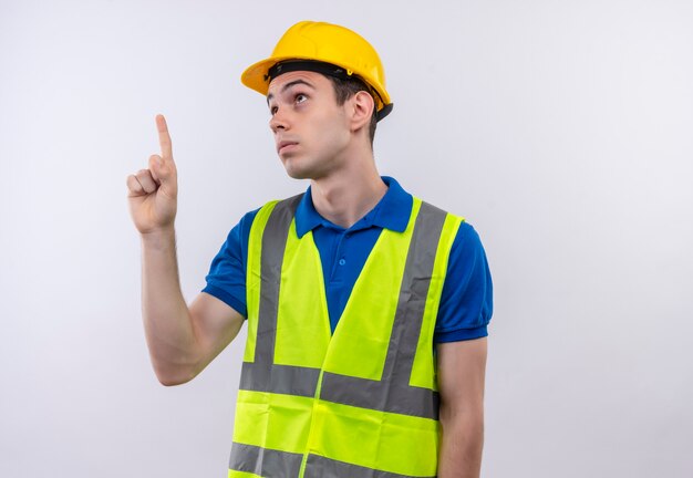 Young builder man wearing construction uniform and safety helmet points up confused