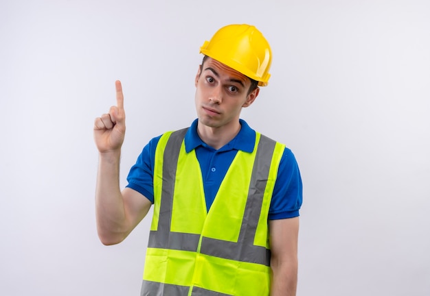 Young builder man wearing construction uniform and safety helmet points up surprised