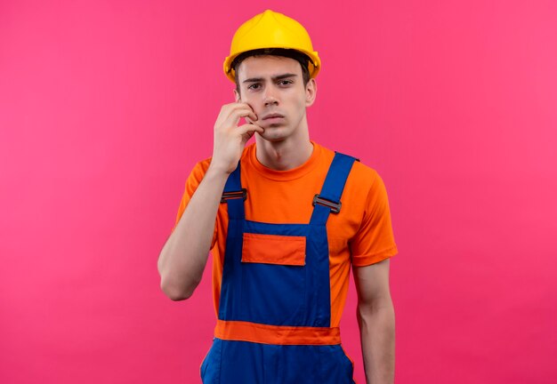 Молодой строитель в строительной форме и защитном шлеме трет лицо левой рукой