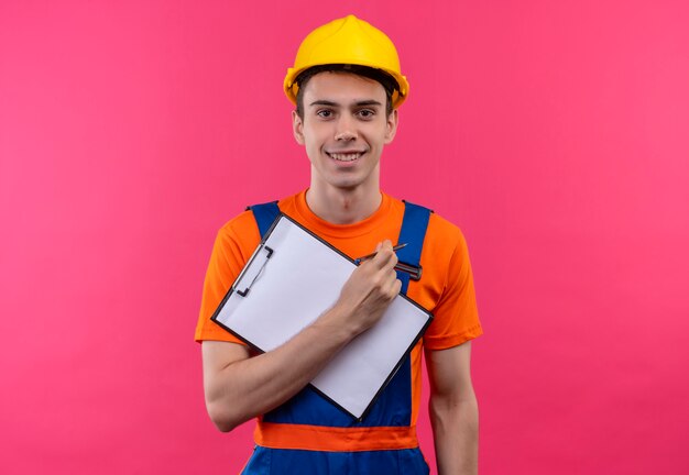 Young builder man wearing construction uniform and safety helmet smiles and holds a clipboard and a pen