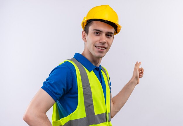 Young builder man wearing construction uniform and safety helmet smiles and shows besides