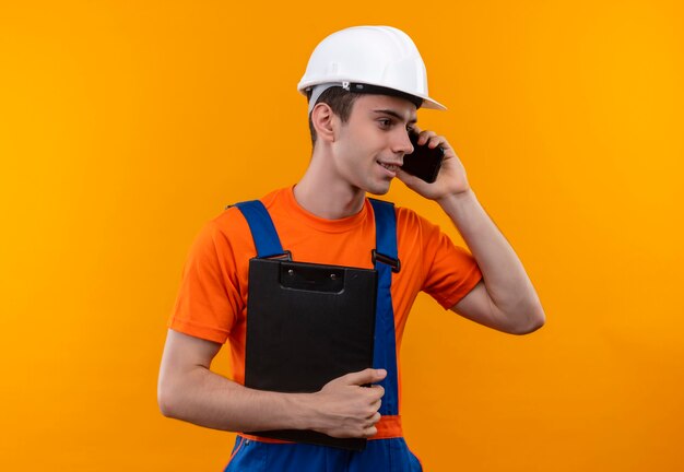 Young builder man wearing construction uniform and safety helmet talks on phone and holds clipboard