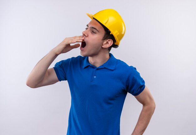 Young builder man wearing construction uniform and safety helmet yawns