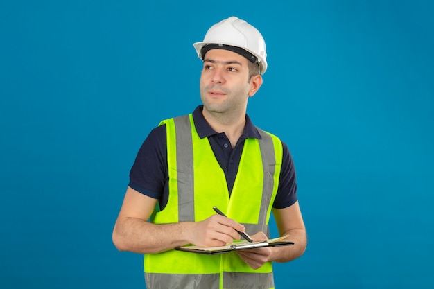 Young builder man wearing white helmet and a yellow vest, looking aside holding clipboard and pen standing on blue isolated