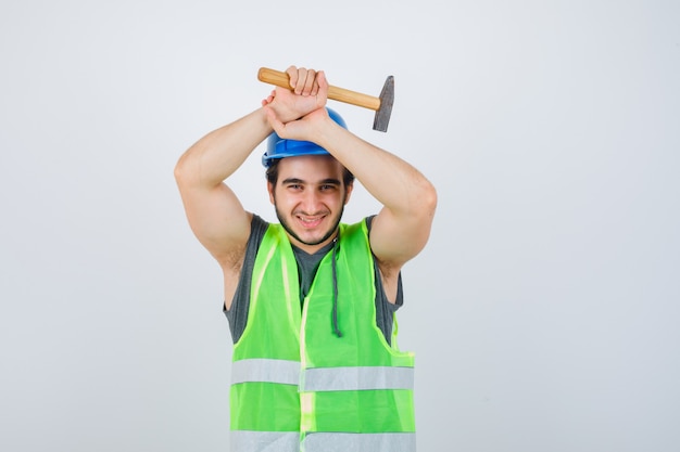 Young builder man in workwear uniform holding hammer over head and looking joyful , front view.