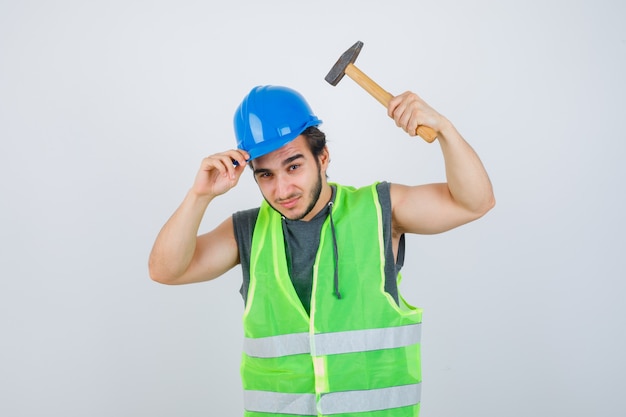 Молодой строитель человек в форме спецодежды держит шлем и молот и выглядит уверенно, вид спереди.
