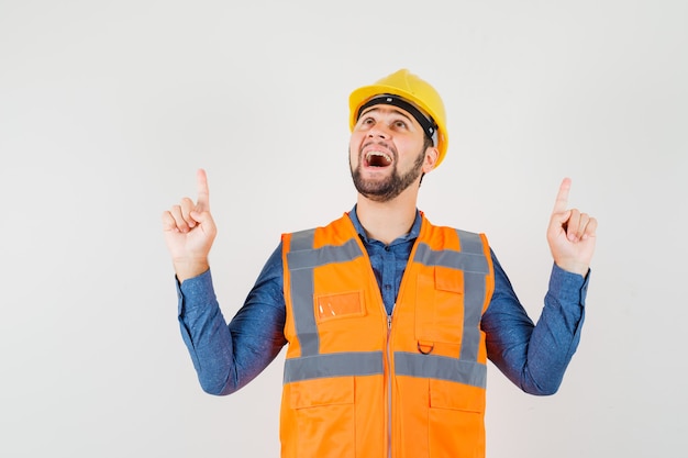 Young builder pointing fingers up in shirt, vest, helmet and looking grateful , front view.