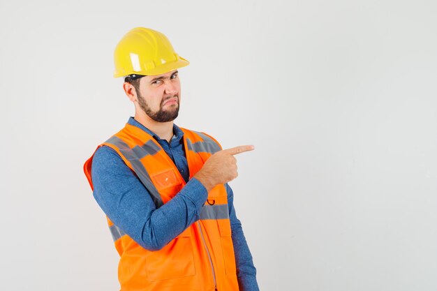 Young builder pointing to the side in shirt, vest, helmet and looking disgusted , front view.