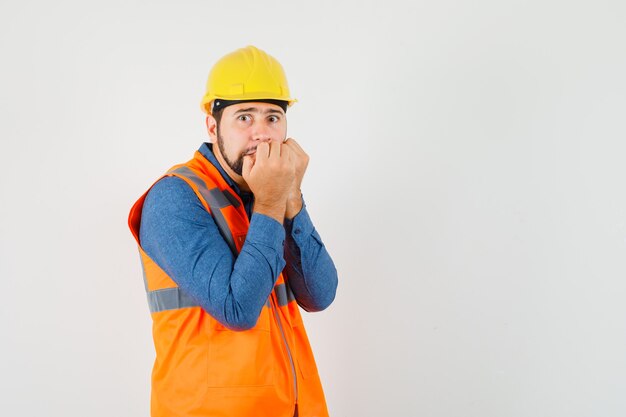 Young builder in shirt, vest, helmet biting fists emotionally and looking frightened , front view.