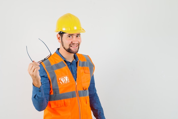 Young builder in shirt, vest, helmet holding glasses and looking cheerful , front view.