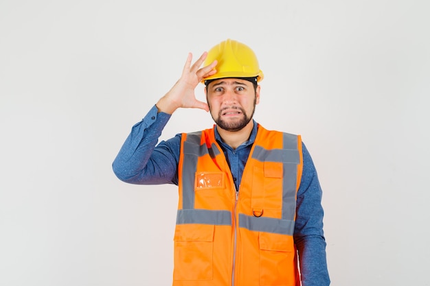 Young builder in shirt, vest, helmet holding hand over head and looking mournful , front view.