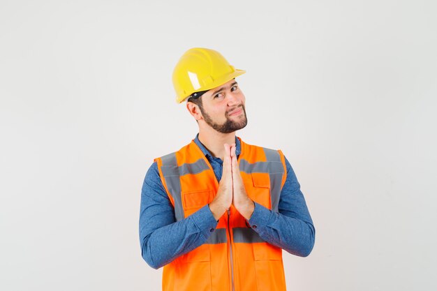 Young builder in shirt, vest, helmet holding hands in praying gesture and looking optimistic , front view.