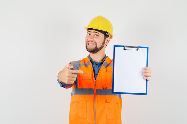 Young builder in shirt, vest, helmet pointing at clipboard and looking cheerful , front view.