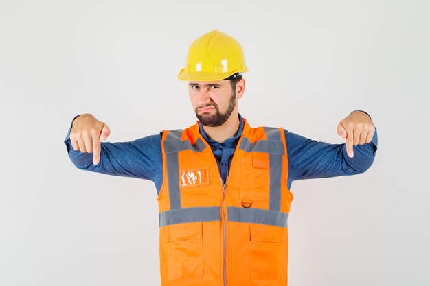 Young builder in shirt, vest, helmet pointing fingers down and looking discontent , front view.