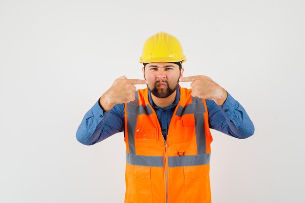 Young builder in shirt, vest, helmet pointing at his nose while frowning , front view.