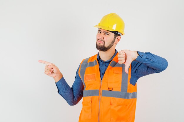 Young builder in shirt, vest, helmet showing thumb down, pointing to the side and looking discontent , front view.