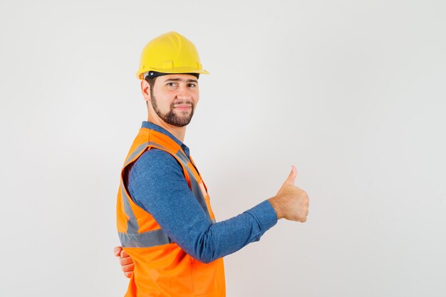 Young builder in shirt, vest, helmet showing thumb up and looking pleased .