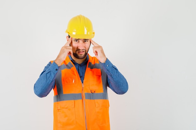 Young builder in shirt, vest, helmet suffering from strong headache and looking tired , front view.