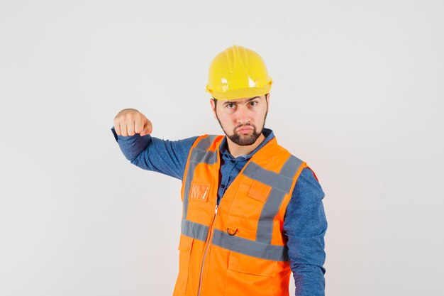 Young builder in shirt, vest, helmet threatening with fist and looking serious , front view.