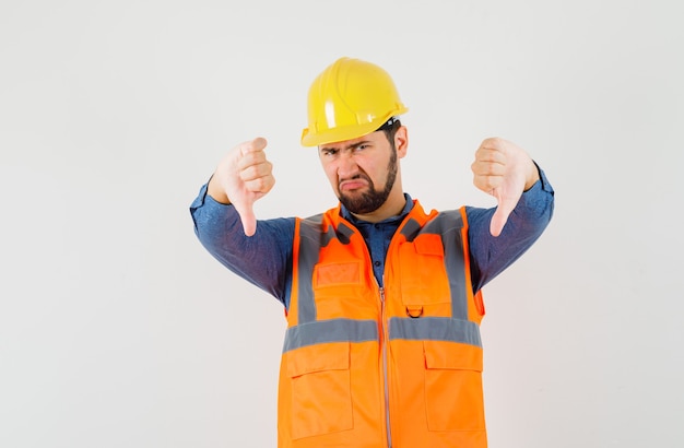 Молодой строитель показывает двойные пальцы вниз в рубашке, жилете, шлеме и выглядит разочарованным, вид спереди.