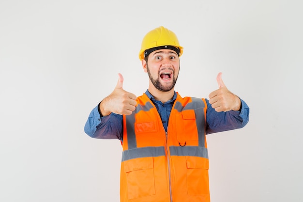 Молодой строитель показывает двойные пальцы вверх в рубашке, жилете, шлеме и выглядит удачливым, вид спереди.