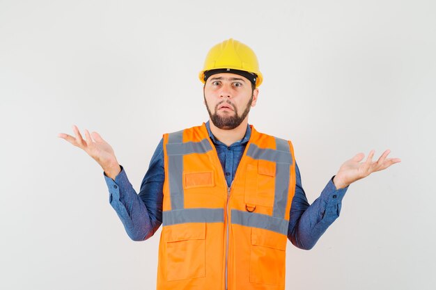 Giovane costruttore che mostra gesto impotente in camicia, gilet, casco e che sembra confuso. vista frontale.