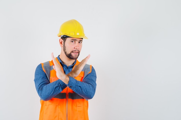 Молодой строитель показывает жест остановки в рубашке, жилете, шлеме и выглядит раздраженным. передний план.