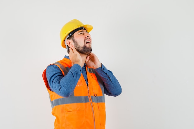 Giovane costruttore che soffre di dolore al collo in camicia, gilet, casco e sembra esausto, vista frontale.