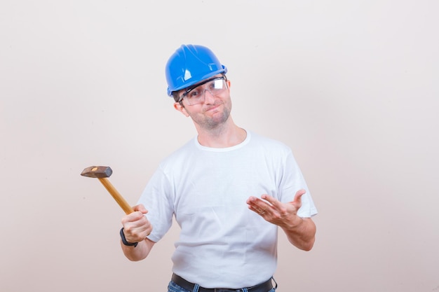Young builder in t-shirt, jeans, helmet showing hammer and looking disappointed