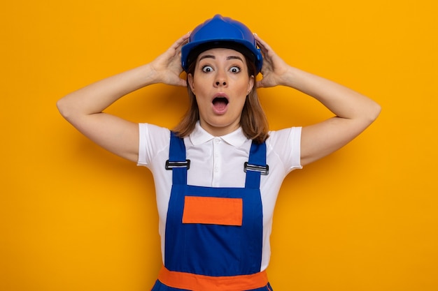 Young builder woman in construction uniform and safety helmet  amazed and surprised with hands on her head standing over orange wall