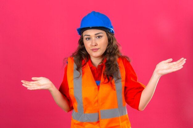 Young builder woman in construction uniform and safety helmet clueless and confused expression with arms and hands raised doubt concept over isolated pink wall