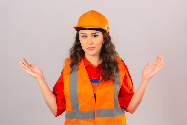 Young builder woman in construction uniform and safety helmet clueless and confused expression with arms and hands raised over isolated white wall
